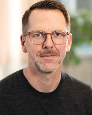 Ein Mann mit Brille und einem kurzen Bart trägt ein schwarzes T-Shirt. Er schaut direkt in die Kamera, mit einem neutralen Gesichtsausdruck. Im Hintergrund sind unscharfe, helle Farben und Pflanzen zu sehen.