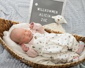 Neugeborenes Baby in einem Korb, trägt einen weißen Overall mit Blumenmuster. Neben dem Baby steht ein Schild mit "Willkommen Alicia Zoe".