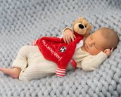 Ein schlafendes Baby in einem weißen Strampler, das ein rotes Kuscheltuch mit einem Bären hält, liegt auf einer grauen, gestrickten Decke.