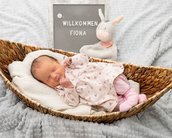 Neugeborenes Mädchen schläft in einem Korb, umgeben von einer Decke und einem Stoffhasen. Ein Schild mit "Willkommen Fiona" steht daneben.
