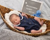 Neugeborenes Baby in einem Korb, trägt einen blauen Overall. Neben ihm steht ein Schild mit "Willkommen Henry Artur" und einem blauen Spielzeug.