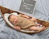 Ein schlafendes Baby in einem Korb, gekleidet in einem braunen Strickanzug, mit einem Schild, das "Willkommen Emre Oskar" zeigt.