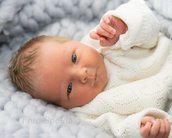 Neugeborenes Baby mit blauen Augen und hellem Haar, gekleidet in einen weißen Pullover, liegt auf einer weichen, grauen Decke.