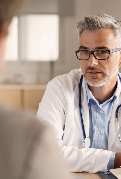 Ein Arzt mit Brille und Stethoskop sitzt an einem Tisch und spricht mit einem Patienten. Der Arzt hält einen Stift und hat ein Notizbuch vor sich. Der Patient ist von hinten sichtbar.