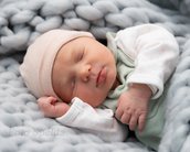 Ein schlafendes Neugeborenes in einem weichen, grauen Strickmuster, trägt einen rosa Hut und einen grünen Schlafanzug. Das Baby liegt auf einer grauen Decke und hat eine entspannte Körperhaltung.