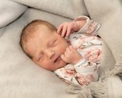 Neugeborenes Baby schläft friedlich in einer Decke, trägt einen weißen Body mit Blumenmuster und hat ein Armband am Handgelenk.