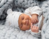 Ein schlafendes Baby in einem weichen, grauen Strickbett, trägt einen weißen Hut und einen bodenlangen Schlafanzug mit Tiermotiven.
