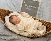 Neugeborenes Baby in einem Korb, trägt einen hellen Strickanzug. Ein Schild mit der Aufschrift "Willkommen Mila" steht daneben.