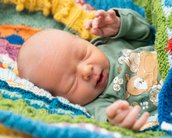 Ein schlafendes Baby in einem bunten, gestrickten Decken, trägt ein grünes Oberteil mit Tiermotiven und hat die Hände leicht erhoben.