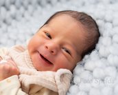Ein lächelndes Neugeborenes liegt auf einer weichen, grauen Decke. Das Baby trägt einen hellen Overall und hat dunkles Haar.