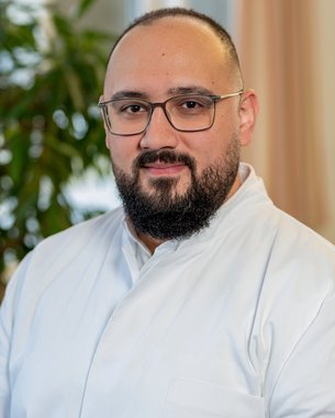 Ein Mann mit Brille und Bart trägt einen weißen Kittel. Er steht vor einem Fenster mit Pflanzen im Hintergrund.
