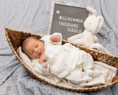 Neugeborenes Baby in einem Korb, umgeben von einer Decke, mit einem Schild, das "Willkommen Theodore Tassilo" zeigt, und einem Plüschhasen.