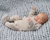 Ein Baby liegt auf einer grauen, gestrickten Decke und trägt einen beigen Strampler. Es hat die Hände erhoben und schaut neugierig.