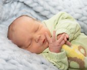 Neugeborenes Baby schläft friedlich auf einer weichen Decke, trägt einen grünen Overall mit einem bunten Muster und hat die Hände zusammengelegt.