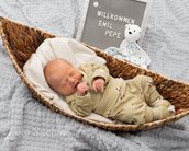 Neugeborenes Baby in einem Korb, trägt einen beigen Schlafanzug. Neben ihm steht ein Schild mit der Aufschrift „Willkommen Emmi Pepe“.