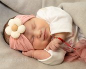 Schlafendes Baby mit einem rosa Haarband, verziert mit einer Blume, trägt ein weißes Oberteil und hat eine rote Armbänder.