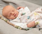 Schlafendes Baby in einem gestreiften Overall mit Tiermotiven, auf einer grauen Decke liegend, umgeben von Holzperlen.
