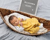 Neugeborenes Baby in einem geflochtenen Korb, gekleidet in einem gelben Overall, liegt auf einer weichen Decke. Ein Schild im Hintergrund zeigt den Namen "Georg" und den Willkommensgruß.