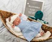 Neugeborenes Mädchen in einem Korb, trägt ein blaues Kleid und einen Haarreifen. Neben ihr steht ein Schild mit ihrem Namen und Geburtsdaten.