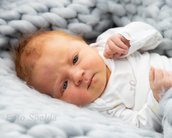 Ein Neugeborenes mit blauen Augen und leichtem Lächeln liegt auf einer grauen, gestrickten Decke. Es trägt einen weißen Bodysuit.