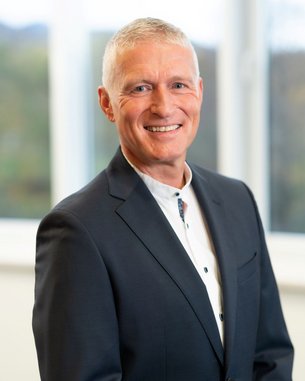 Ein Mann mit kurzen, grauen Haaren und einem freundlichen Lächeln trägt einen dunklen Anzug und ein helles Hemd. Er steht vor einem Fenster mit unscharfem Hintergrund.