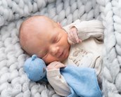 Neugeborenes Baby schläft friedlich in einer grauen Decke, umarmt ein blaues Kuscheltier und trägt einen hellen Strampler.