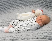Neugeborenes Baby schläft auf einer grauen Decke, trägt einen weißen Overall mit schwarzen Punkten und hat ein gehäkeltes Spielzeug neben sich.