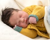 Ein schlafendes Neugeborenes in einem gelben Strickpullover mit grünen Akzenten, eingekuschelt in eine weiche Decke.