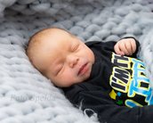 Ein schlafendes Baby liegt auf einer weichen, grauen Decke, trägt ein schwarzes Oberteil mit buntem Aufdruck und hat eine entspannte Körperhaltung.