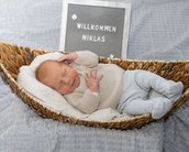 Neugeborenes Baby liegt in einem Korb, trägt einen hellen Pullover und gestreifte Hosen. Ein Schild im Hintergrund zeigt "Willkommen Niklas".